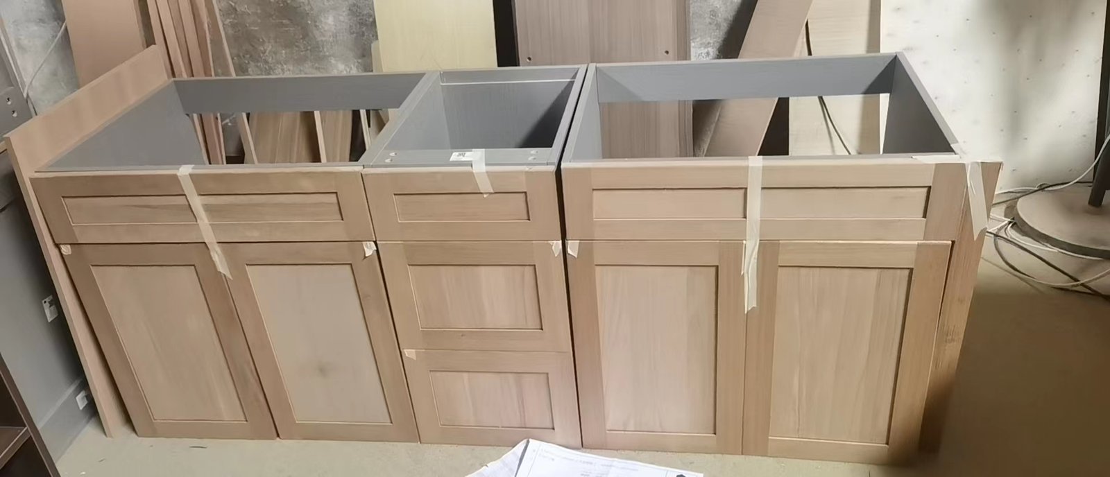 Three light wood kitchen base cabinet assemblies with framed door fronts and drawers in factory workshop
