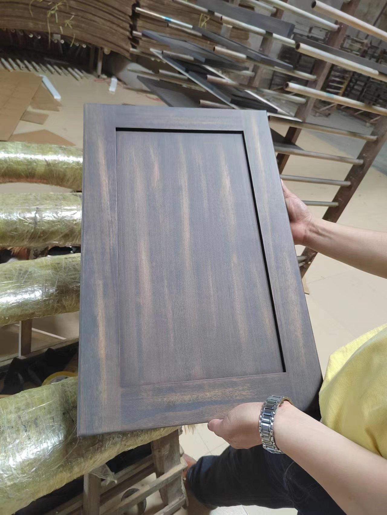 Worker holding a dark stained wood cabinet door sample in a woodworking workshop