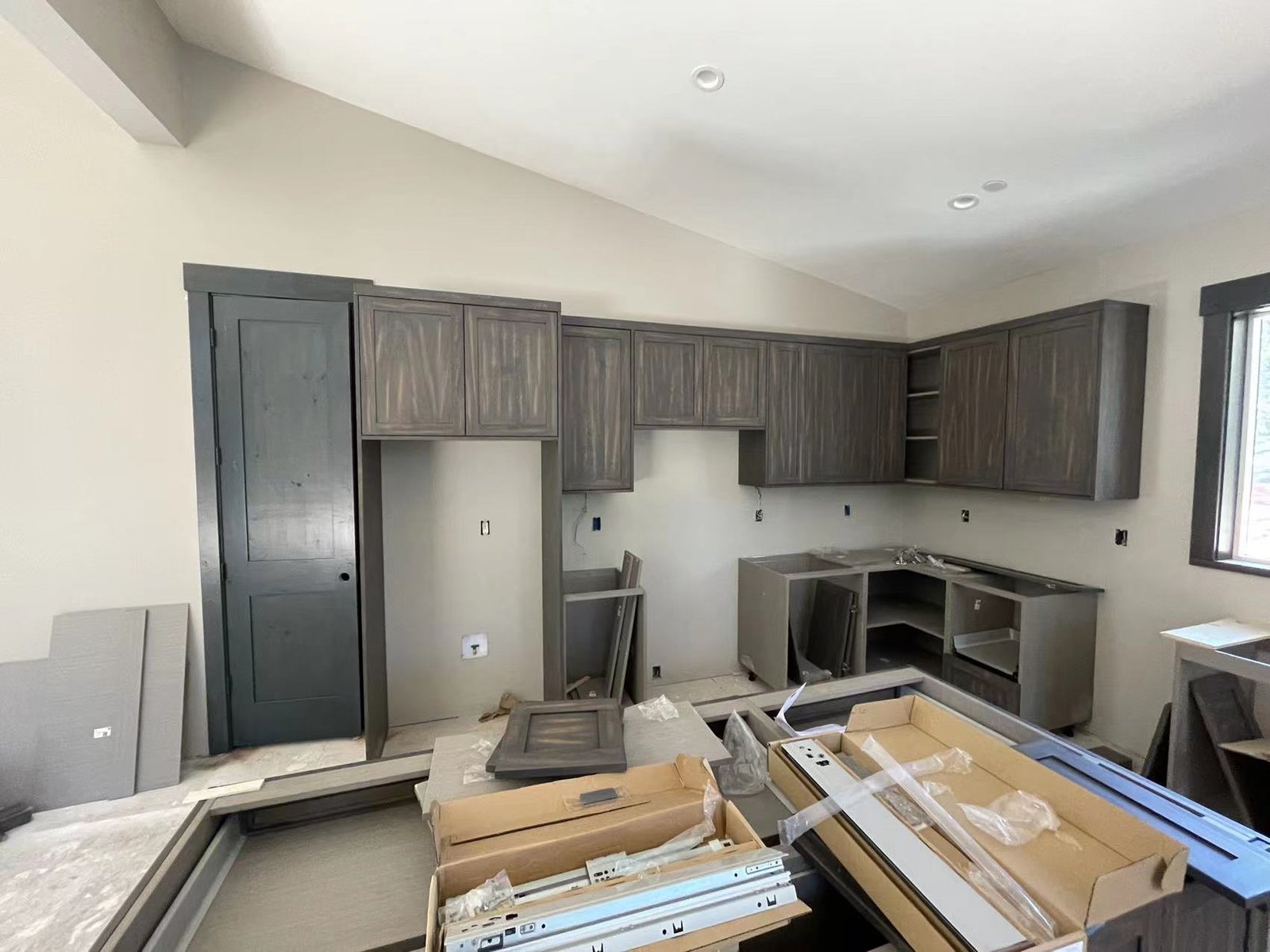 Partially installed dark wood kitchen cabinets at a residential construction site, with hardware and packaging in foreground