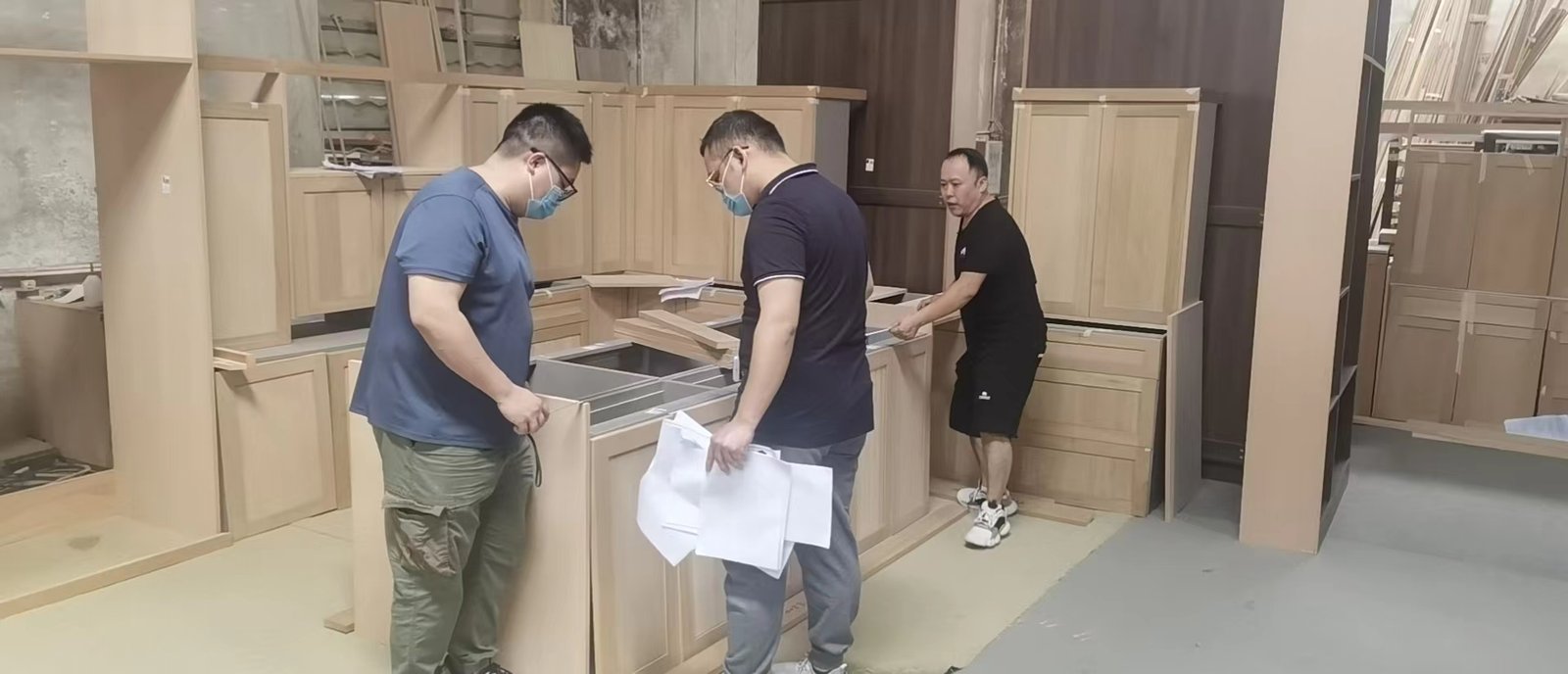 Three people reviewing printed technical drawings next to unfinished kitchen cabinets in a manufacturing workshop